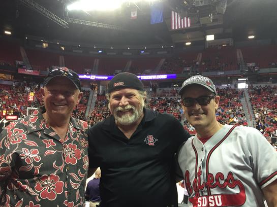 Three friends smile for the camera while attending a vibrant baseball game with excited fans.
