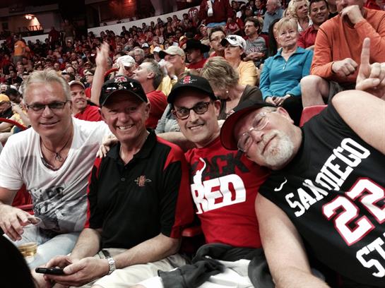 A group of friends sits together, smiling and cheering, wearing team gear in a vibrant crowd.