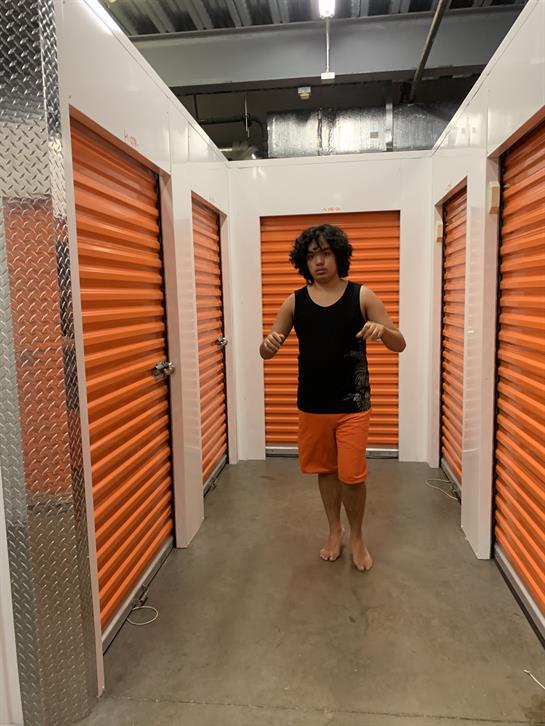A young man in casual attire walks barefoot through a storage facility with orange doors.