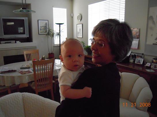 A grandmother lovingly holds her baby grandchild in a sunny living room filled with warmth.