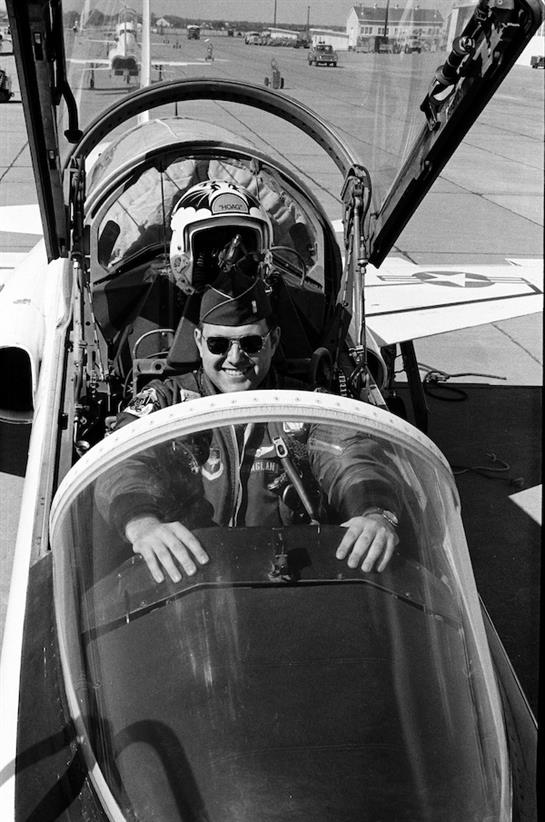A smiling pilot sits in the cockpit of a military plane, ready for takeoff on a clear day.