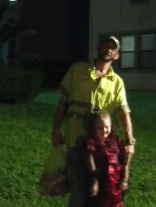 A man stands behind a smiling child in casual attire outside at night with green grass.