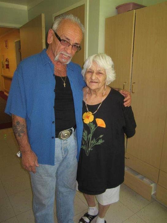 Two older adults smile and embrace while standing in a well-lit hallway, enjoying a joyful moment.