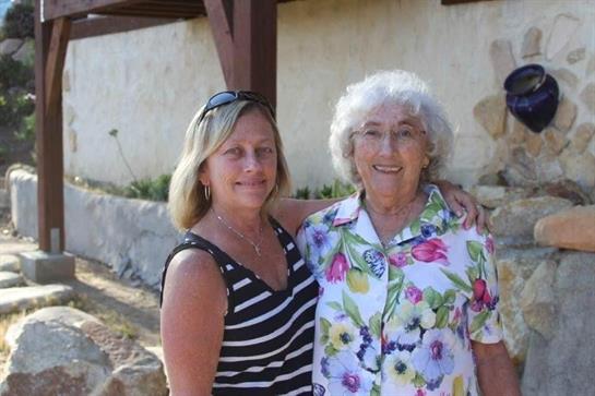 Two women smile and embrace outdoors, surrounded by greenery and a rustic backdrop.