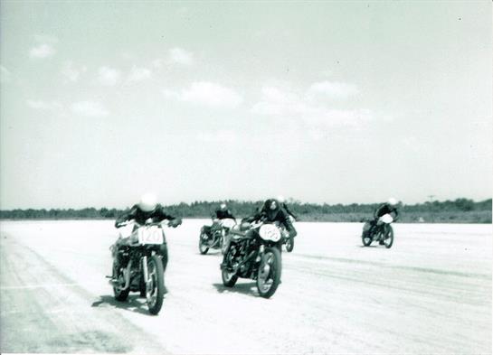 Groups of motorcyclists compete fiercely on a wide, smooth race track surrounded by trees.