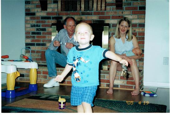 A young boy in a blue shirt and shorts is happily playing while his parents watch him.
