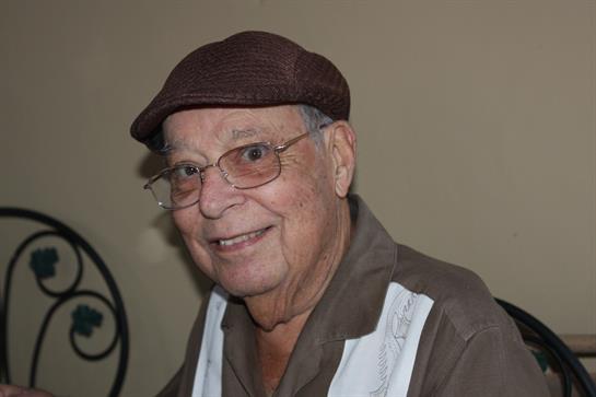 Smiling elderly man in a cap and glasses enjoys indoor company at a casual gathering with friends.