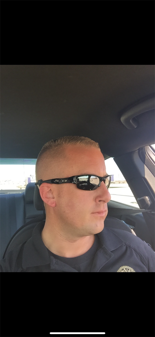A police officer sits inside a patrol vehicle wearing sunglasses, focused on the road ahead.
