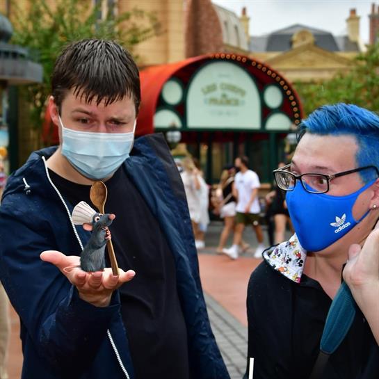 Two people stand outdoors, one holding a toy rat while the other watches, both wearing masks.
