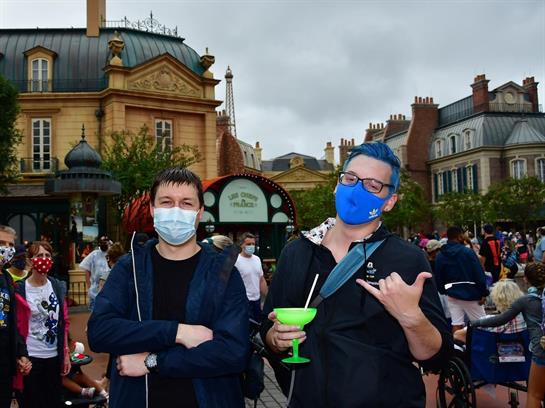 Two friends wearing masks smile at a lively fairground filled with patrons and attractions.