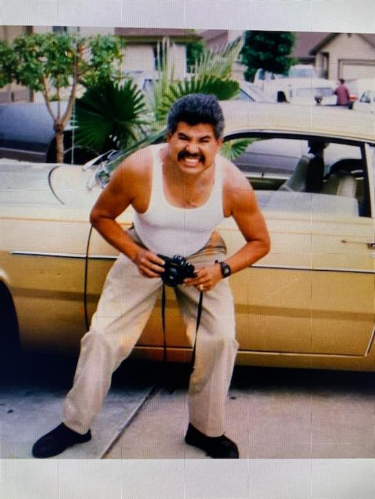 A man in a tank top stands next to a yellow vintage car, holding a camera and smiling broadly.