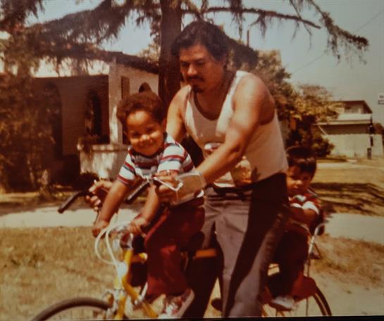 A father rides a bicycle with two kids, enjoying a sunny day in a vibrant neighborhood.