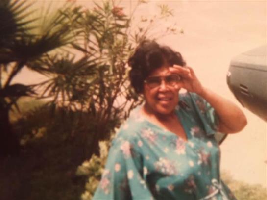 A joyful woman stands outdoors, smiling and shielding her eyes from the sun, surrounded by greenery.