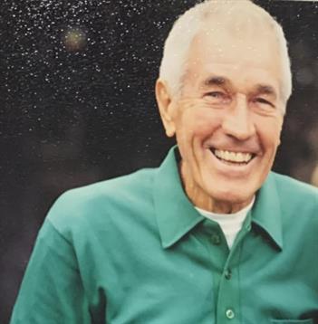 An elderly man with a joyful smile stands outdoors wearing a green shirt under sunlight.