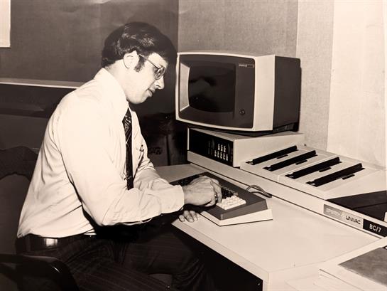 A man types on a keyboard next to a synthesizer and monitor in a retro office during the 1980s.