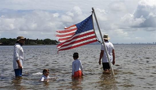 Kids swim and wade in shallow water while an adult holds a faded American flag on a beach.