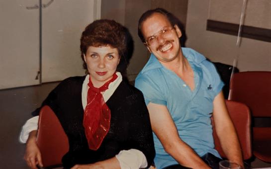 A man and woman sit closely, smiling in casual clothes in a cozy indoor space.