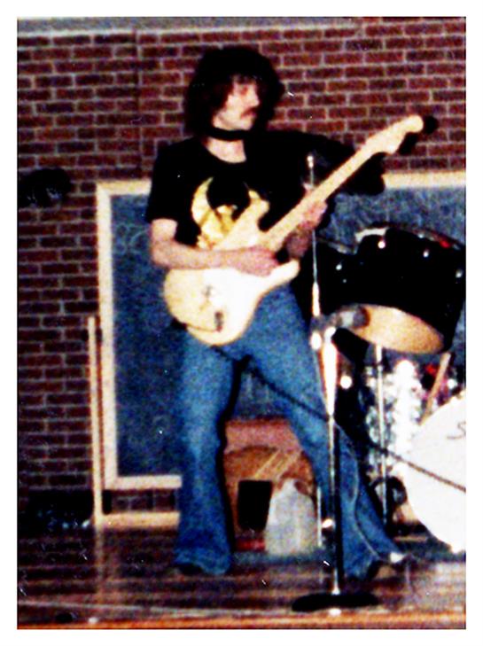 Musician wearing casual clothing plays electric guitar in a school auditorium during a performance.