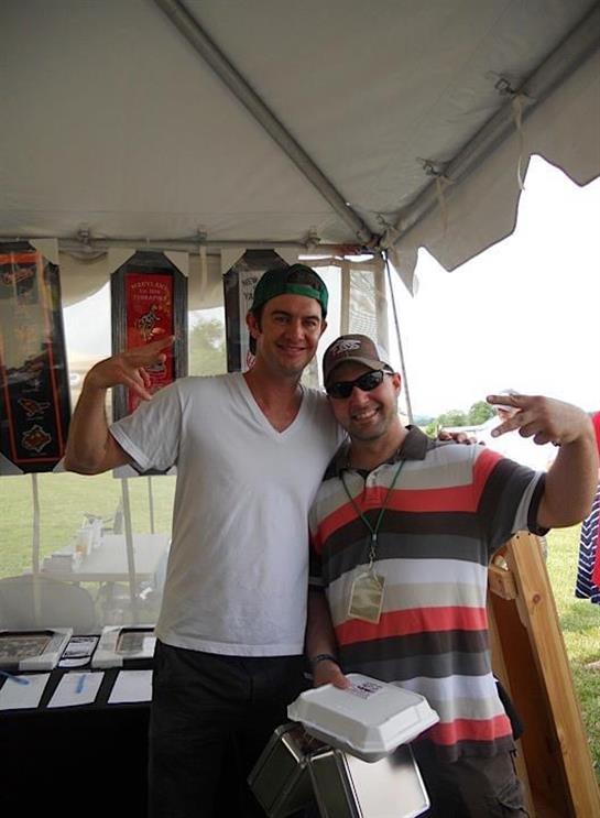 Two men are smiling and making peace signs while enjoying a lively outdoor festival atmosphere.