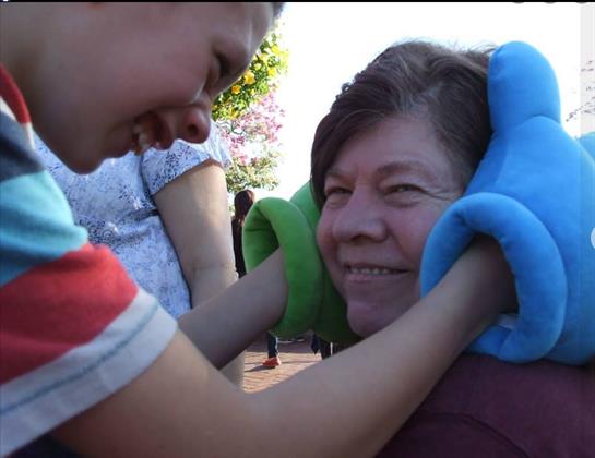 A young person gently teases an elderly woman with colorful gloves on a sunny day in a park.