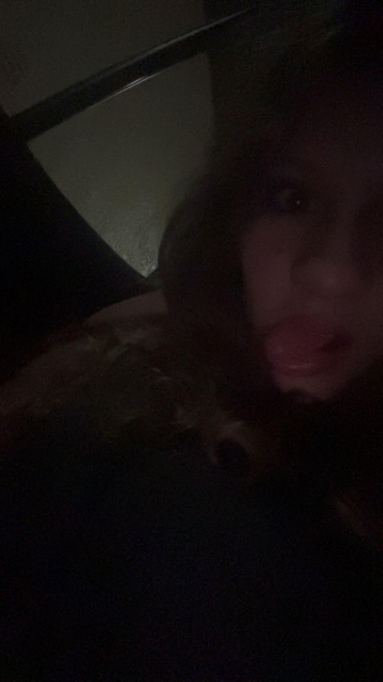 A person is sitting in low light, looking at the camera and showing an expressive face.