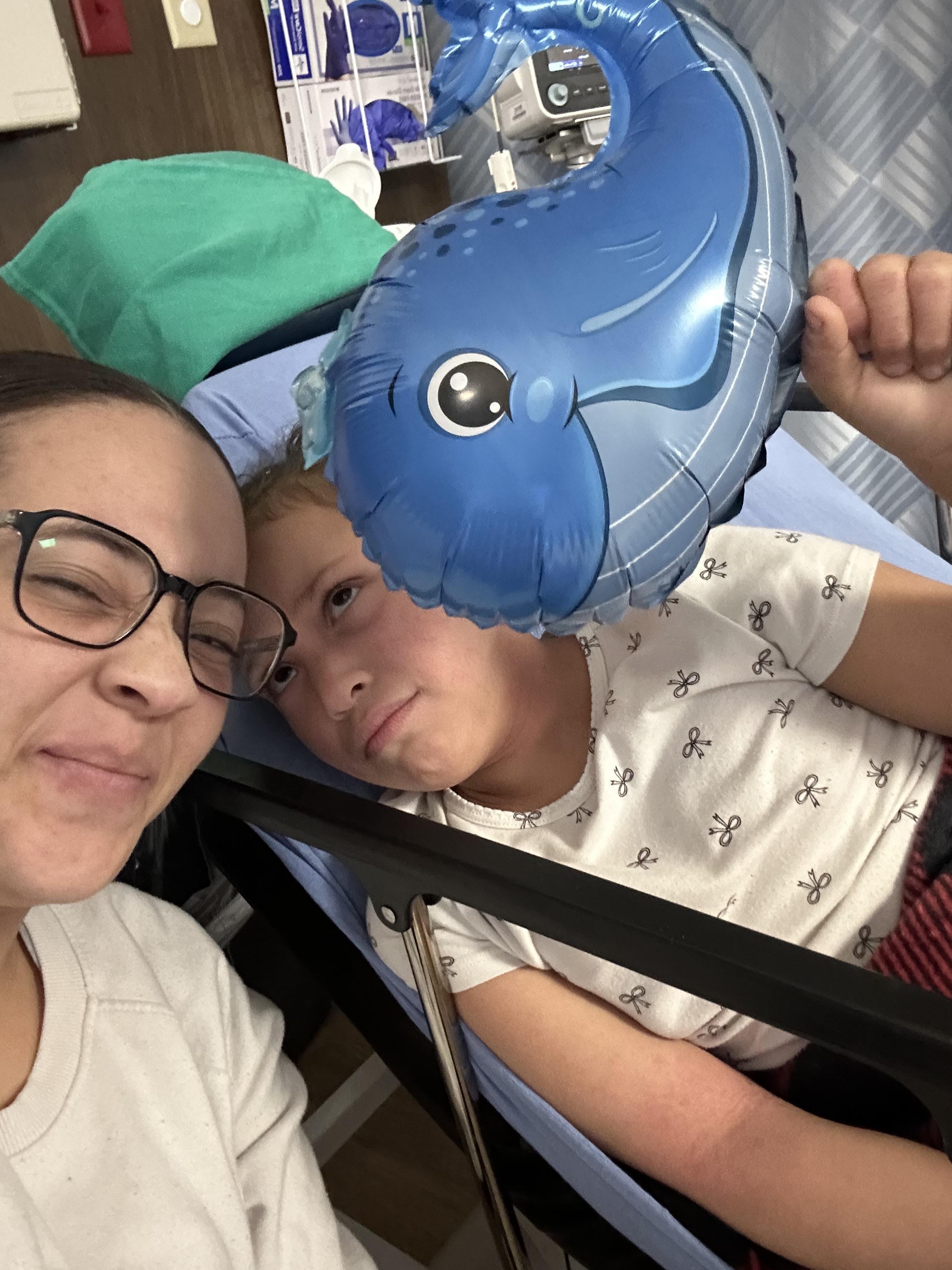 Two people pose for a selfie in a hospital. The child holds a blue balloon.
