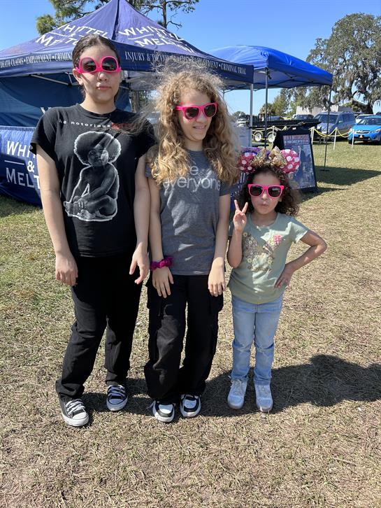 Three children pose together in sunglasses at a lively outdoor gathering on a sunny day.