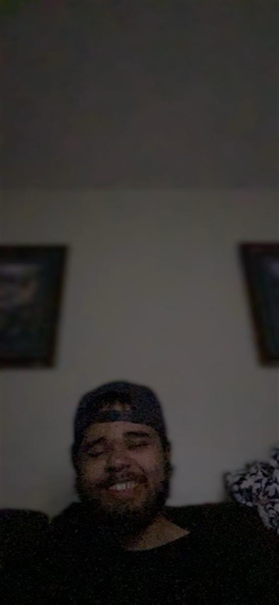 A young man in a cap is sitting in a dark room, talking and engaging with someone nearby.