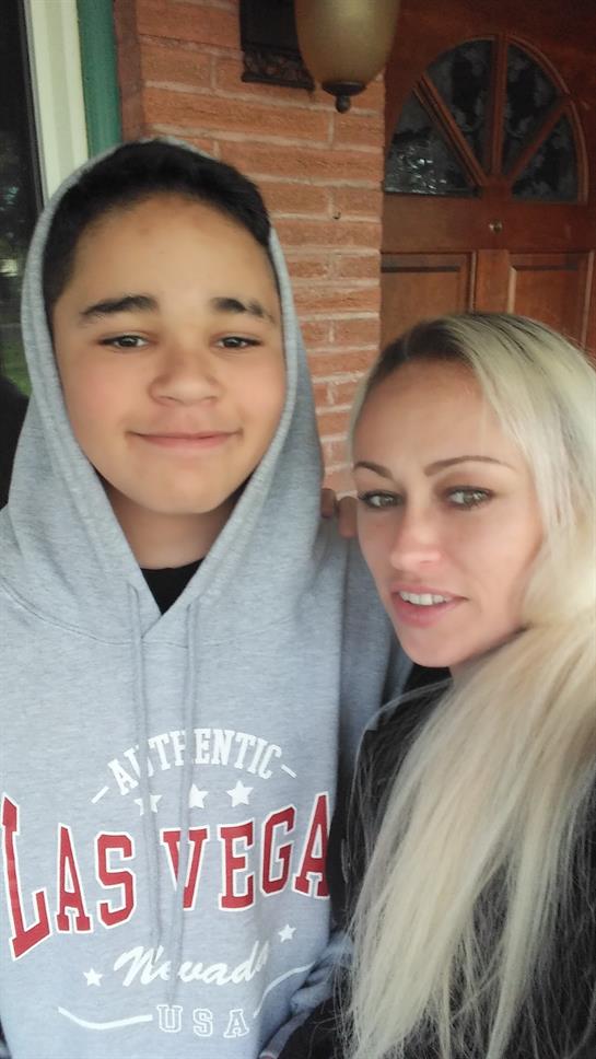 A young boy and his mother stand closely together, smiling in casual clothing, near a brick wall.