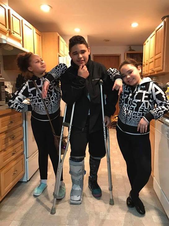 Brother in a cast stands between his smiling sisters in a warmly lit kitchen.