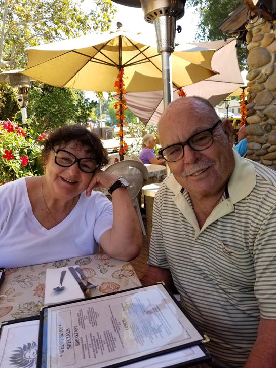 Elderly couple smiles at the camera while dining outdoors on a warm, sunny day.