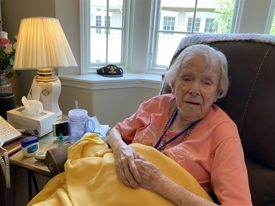 An elderly woman is relaxing in a chair with a warm blanket, enjoying the sunlight in her room.
