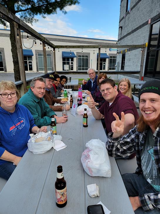 Friends share food and laughter at a long table in a relaxed outdoor setting during the afternoon.
