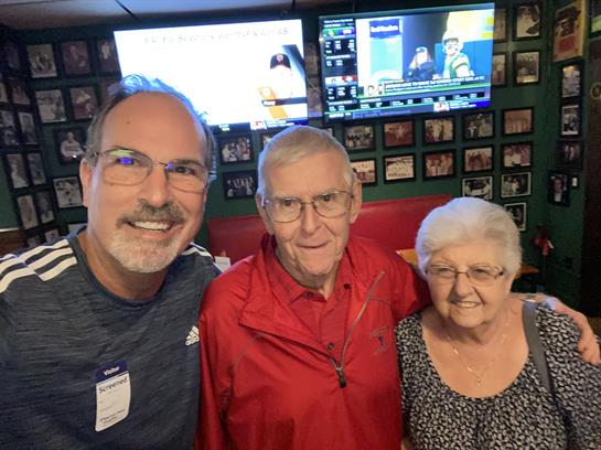 Three people smile while posing together in a lively setting filled with memorabilia.