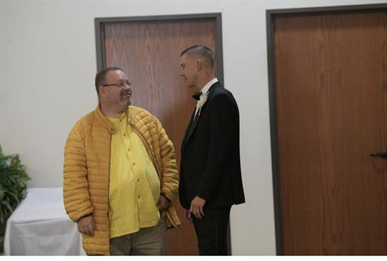 Two men engage in friendly conversation while dressed for a gathering in a simple indoor space.