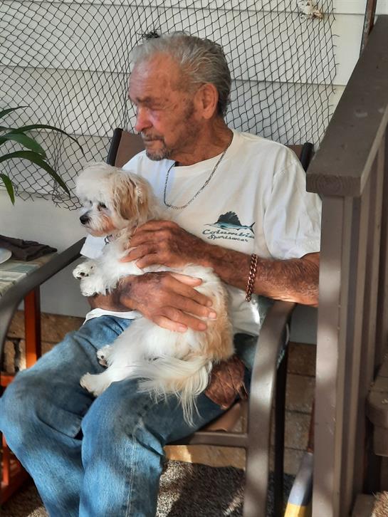An elderly man relaxes on his porch, gently holding a small dog on his lap while enjoying the day.