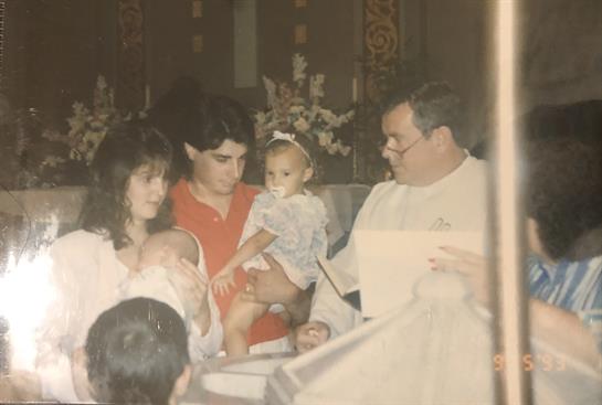 A family participates in a church blessing ceremony with a priest and young child in their arms.