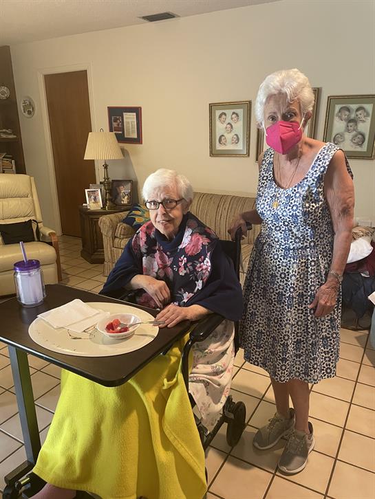 Elderly woman enjoys a snack at a table while caregiver stands nearby in a warm living room setting.