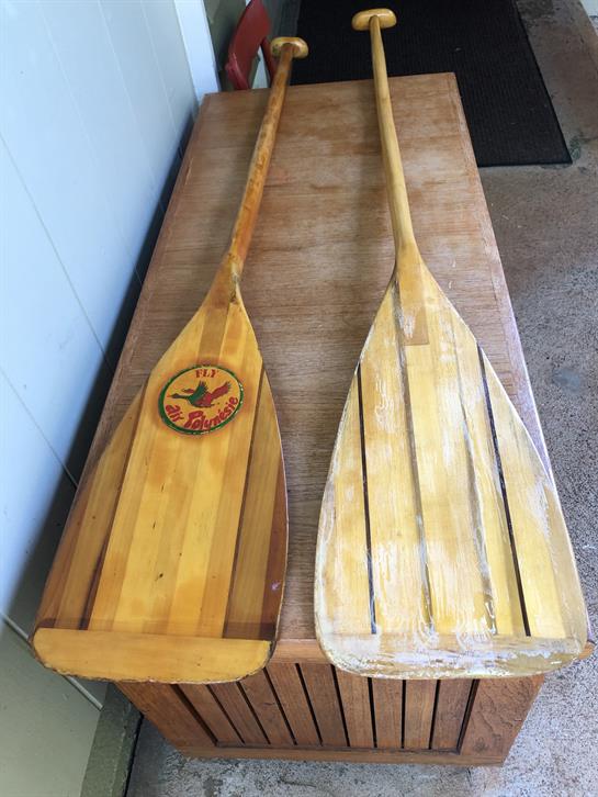 Two rustic wooden paddles rest side by side on a sunlit wooden table outdoors.