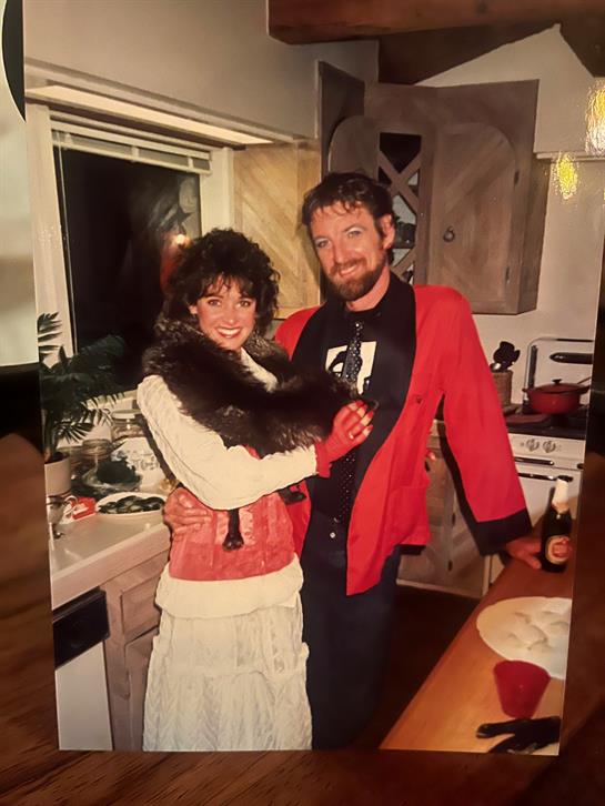 A couple dressed in 1980s fashion share a joyful moment in a kitchen filled with warm light.