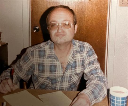 A man in casual attire sits at a table, reviewing papers and sipping from a cup in low light.