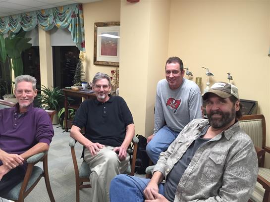 Four men sit comfortably in chairs, engaged in conversation at a relaxed indoor gathering.