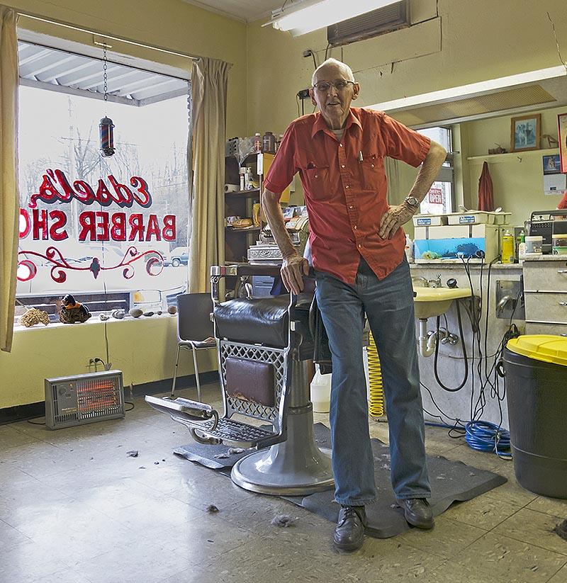 An elderly barber poses confidently in his vintage barbershop filled with tools and chairs.