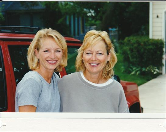 Two women with blonde hair pose happily outdoors beside a red car under clear skies.