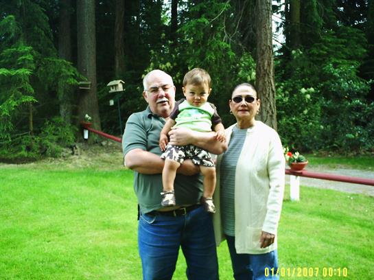 Grandparents smiling while holding their toddler in a green backyard filled with trees.