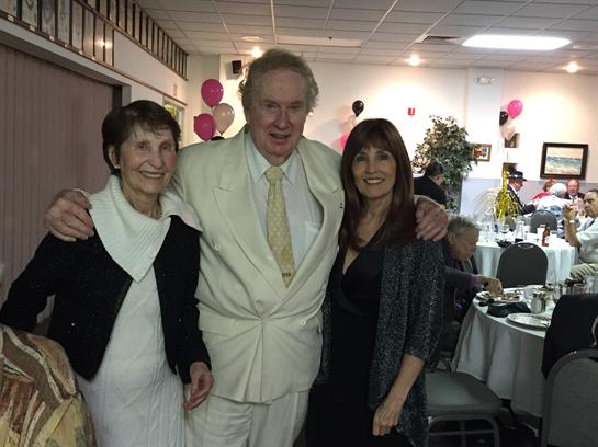 Three individuals are smiling and posing together while dressed elegantly at a festive gathering.