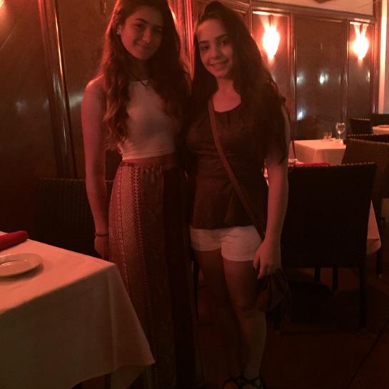 Two young women stand smiling in a warmly lit restaurant, enjoying their night out.