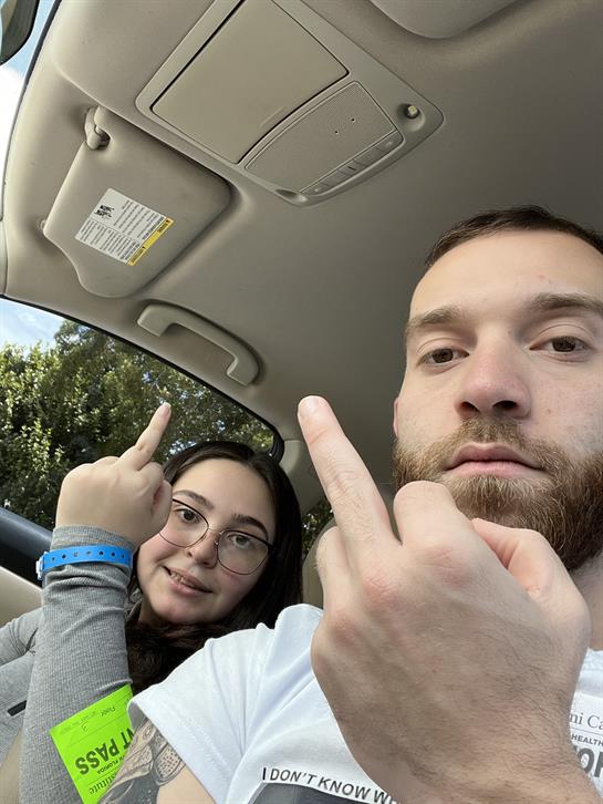 Two friends are seated in a car, playfully showing their middle fingers with smiles.