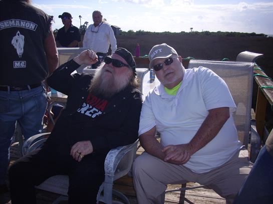 Two men in sunglasses relax in folding chairs at a sunny outdoor event.