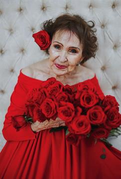 A joyful elderly woman poses gracefully, surrounded by roses while wearing a vibrant red dress.